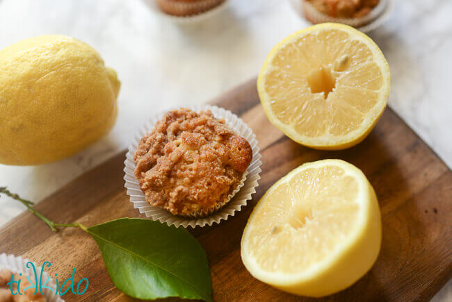Lemon muffin with crumb topping on a wooden cutting board with cut and uncut fresh lemon.
