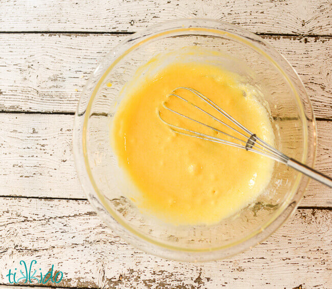 clear pyrex bowl with lemon curd ingredients and whisk, on a weathered white wooden background.