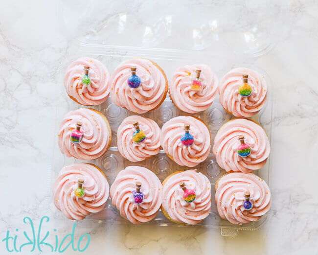 Vanilla cupcakes topped with light pink icing and a tiny bottle full of multi colored sprinkles.