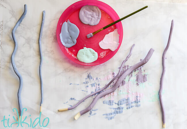Pieces of curly willow being painted in pastel colors, a pink plastic plate with paint and a paintbrush in the center.