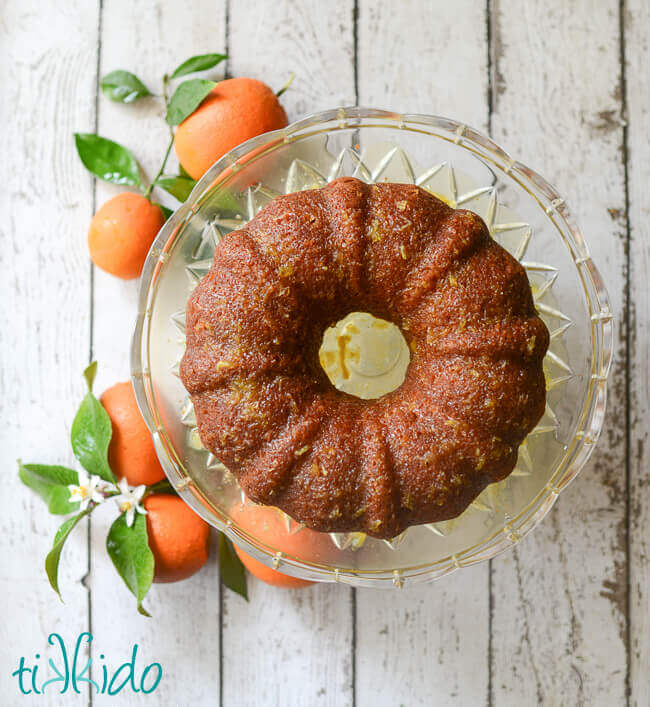 Orange bundt cake with orange glaze on a clear cake stand, surrounded by fresh oranges on the branch.