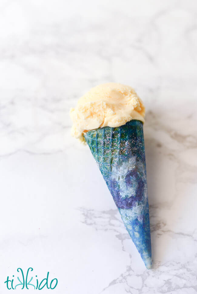 Ice cream cone painted with food coloring like a swirling galaxy with silver disco dust stars, filled with vanilla ice cream.