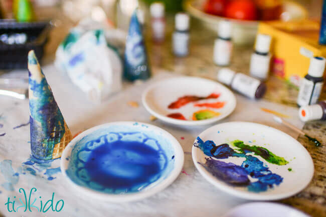 Three small white plates with food coloring paints, bottles of food coloring, and ice cream cones in the process of being painted.