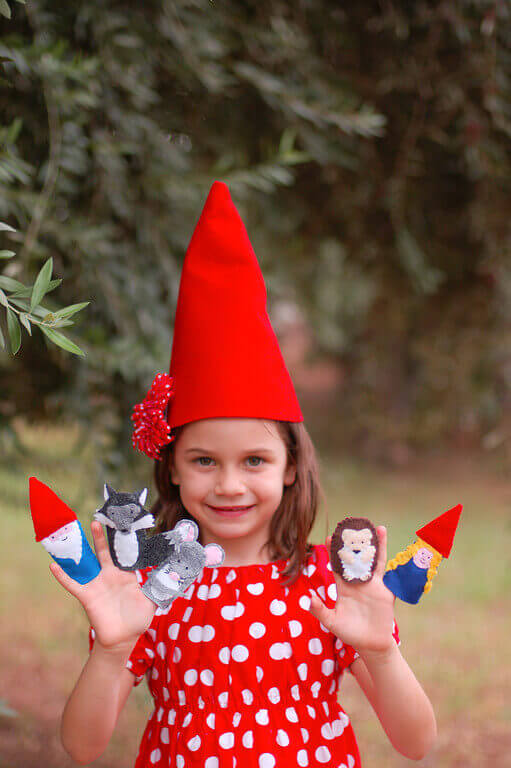 Little girl wearing a gnome hat and a red polka dot dress, with gnome and woodland creature finger puppets on her hands.