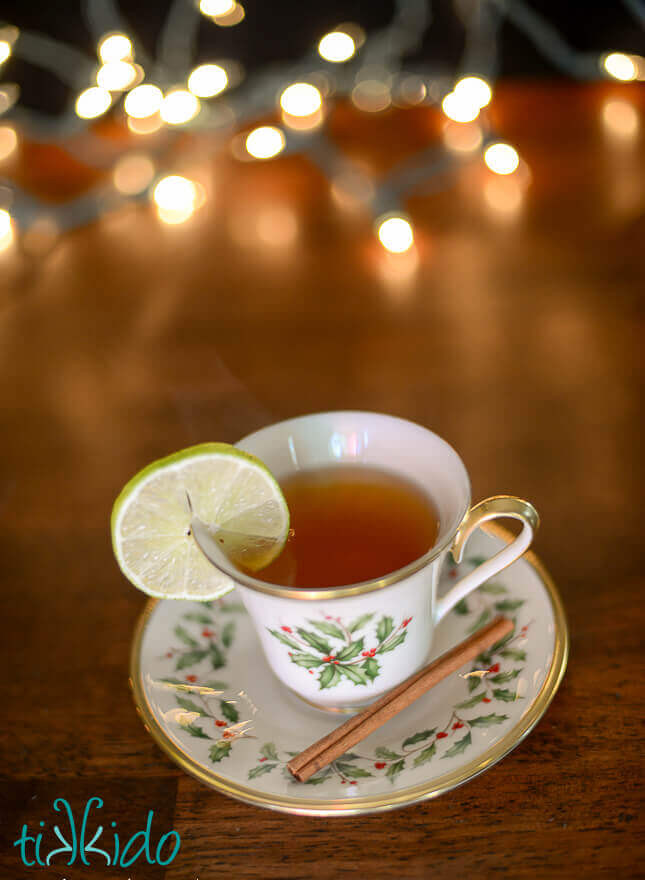 Cup of homemade mulled apple cider in a Christmas teacup, on a matching saucer, with a wedge of lime on the rim of the cup.