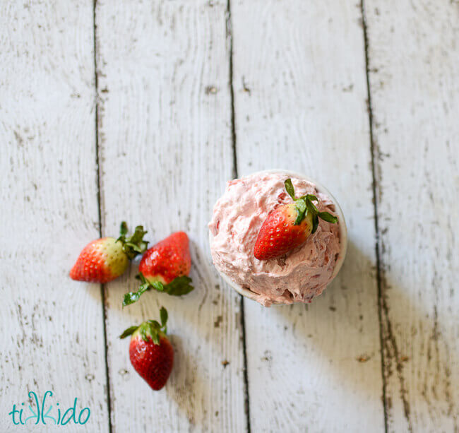 Small white bowl of strawberry cream cheese with a whole strawberry on top, next to three whole strawberries on a white weathered wood surface.
