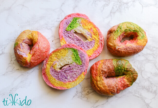 One unicorn bagel cut in half, surrounded by three whole unicorn bagels on a white marble background.