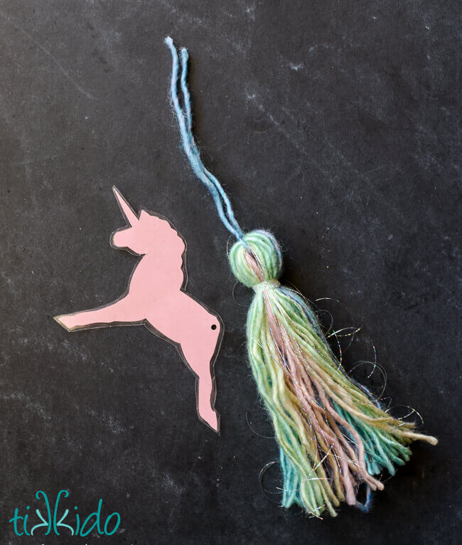 DIY unicorn bookmark with yarn tassel tail being assembled on a black chalkboard background.