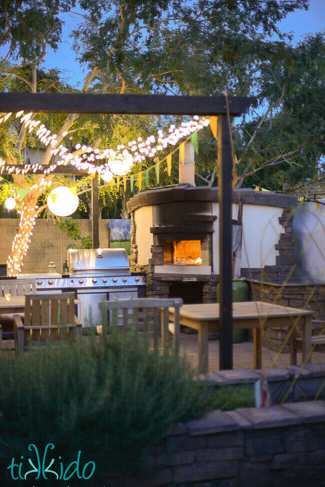 Wood fired pizza oven with a fire inside, framed by an outdoor kitchen and eating area, and covered with a pergola strung with twinkle lights and paper lanterns.
