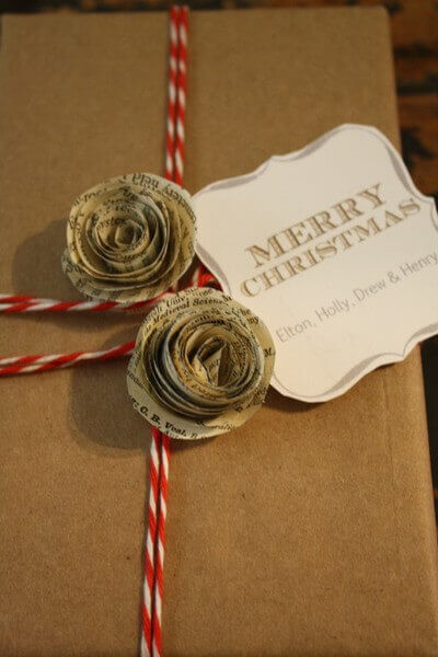 Coiled rose flowers made out of newspaper and red and white string top a plain brown paper wrapped Christmas gift.