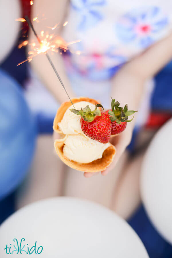 Person holding a waffle cone bowl full of vanilla ice cream and two strawberries, with a lit sparkler stuck in the ice cream.
