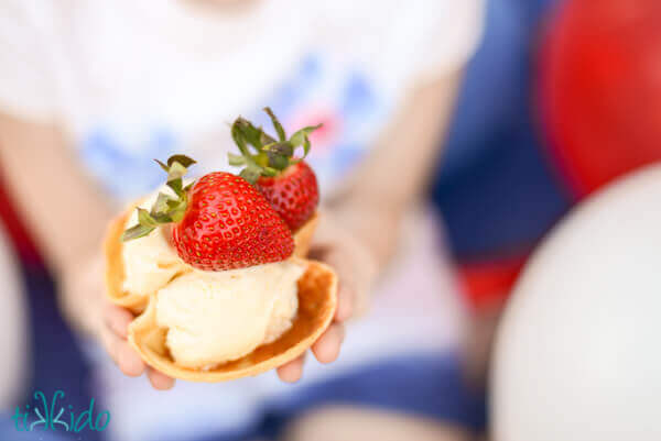 Person holding a waffle cone bowl full of vanilla ice cream and two strawberries, red, white, and blue balloons in the background.