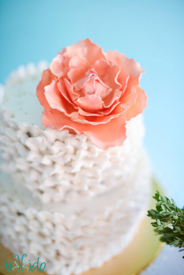 Peach gum paste peony on top of a cake covered in white frosting ruffles.