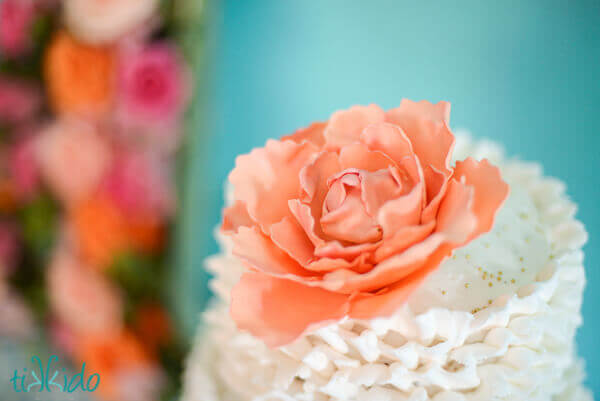 Peach gum paste peony on top of a cake covered in white frosting ruffles.