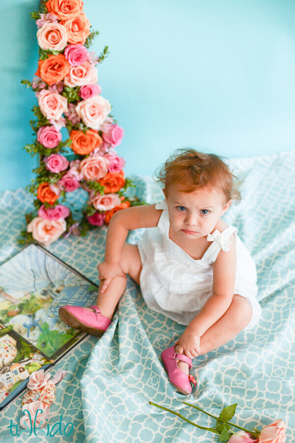 One year old little girl with an alphabet book and a floral letter A, frowning at the camera.