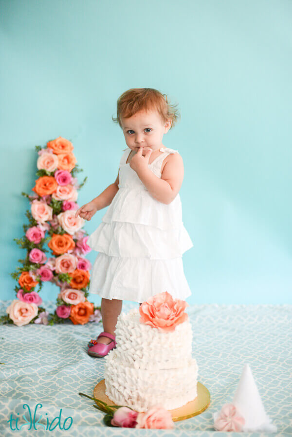 One year old little girl tasting the icing from a two tier cake decorated in white frosting ruffles and a gum paste peony.