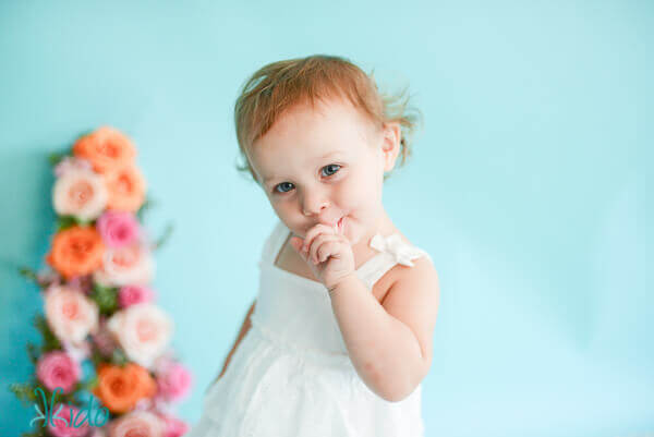 One year old little girl tasting the icing from a two tier cake decorated in white frosting ruffles and a gum paste peony.