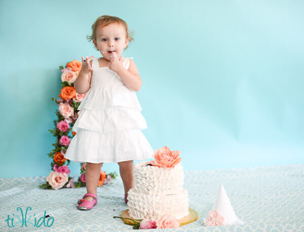 One year old little girl tasting the icing from a two tier cake decorated in white frosting ruffles and a gum paste peony.