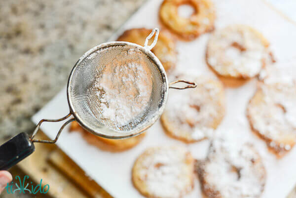 Apple fritter rings being dusted with powdered sugar in a small strainer.