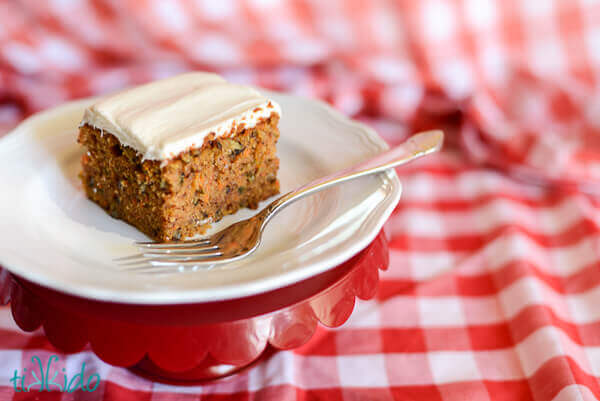 piece of carrot cake made with the best carrot cake recipe, on a white plate, on a red and white checkered tablecloth.