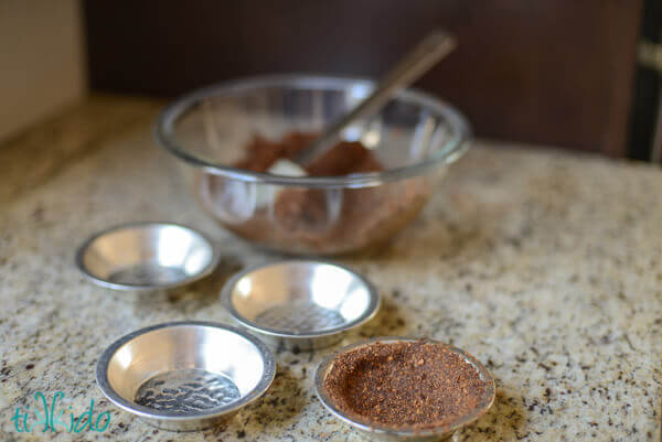 Clear glass bowl of chocolate graham cracker pie crust mixture  and four miniature pie tins, one filled with chocolate graham cracker crust.