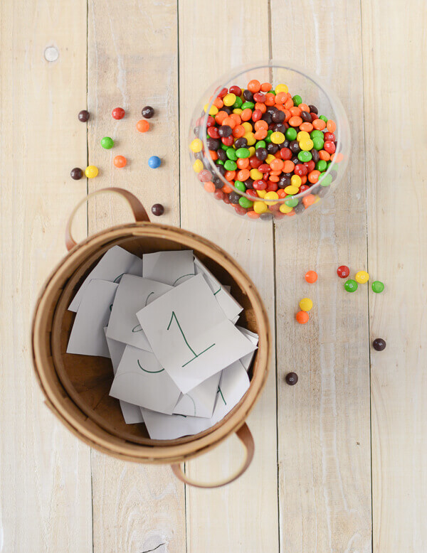 wooden table with a basket full of papers with numbers written on the paper, and a glass bowl full of skittles.