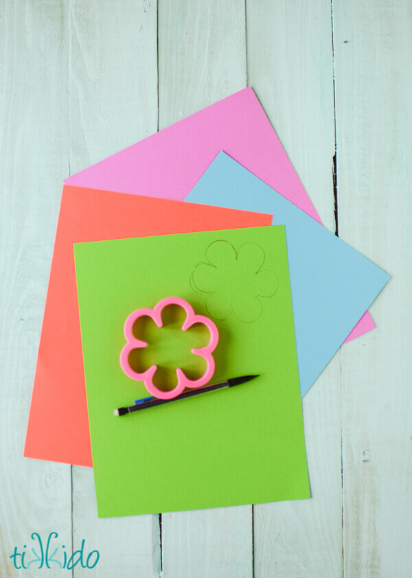 Flower shapes being cut out of colorful cardstock by tracing the shape of a flower cookie cutter.