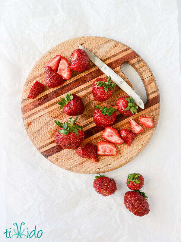 Round wooden cutting board on a white surface, with a knife, and cut and whole strawberries on the board.