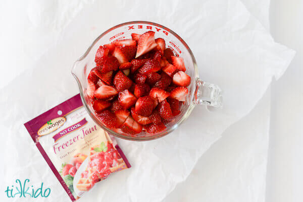 Sliced strawberries in a large pyrex measuring cup, next to a packet of Freezer jam no cook pectin on a white surface.