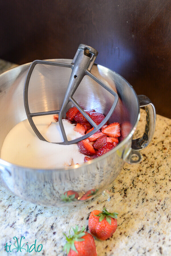 Kitchenaid Mixer bowl with strawberry chunks and sugar and a mixing paddle, on a granite counter.