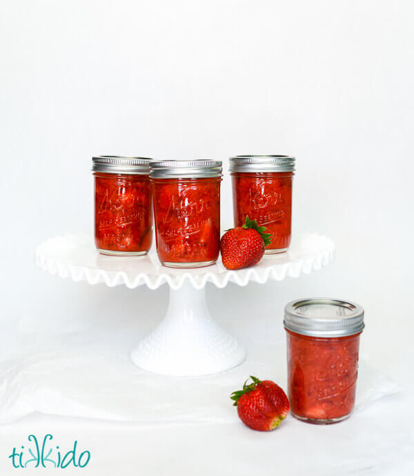 Four jars of Strawberry freezer jam and two fresh strawberries, three jars on a white pedestal, one on the white ground.