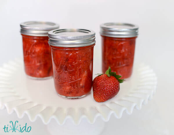 Three jars of strawberry freezer jam and a whole strawberry on a white cake plate on a white background.