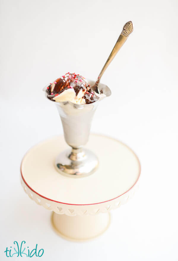 hot fudge sauce on vanilla ice cream, topped with sprinkles, in a silver ice cream dish.