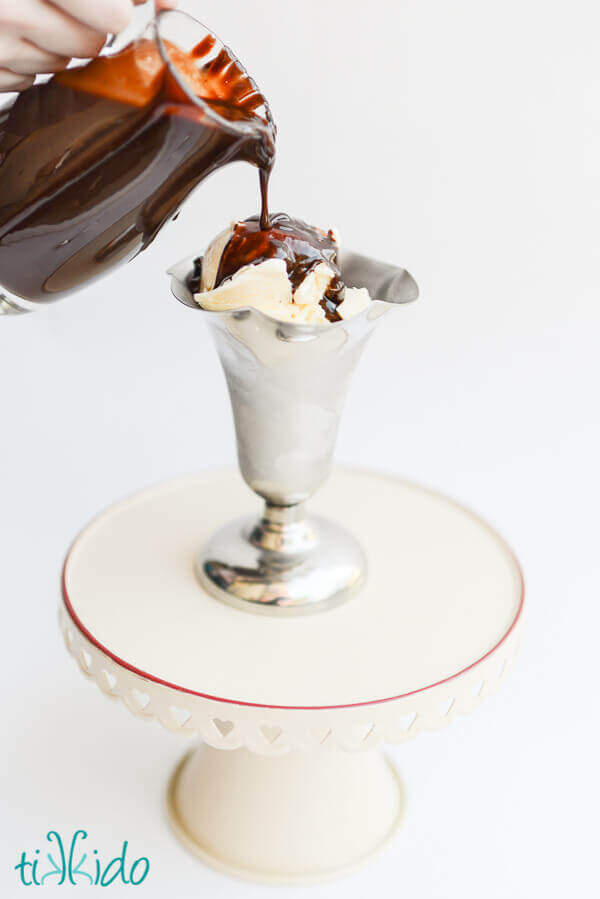 Hot fudge ice cream topping pouring from a pitcher onto a silver ice cream dish filled with vanilla ice cream.