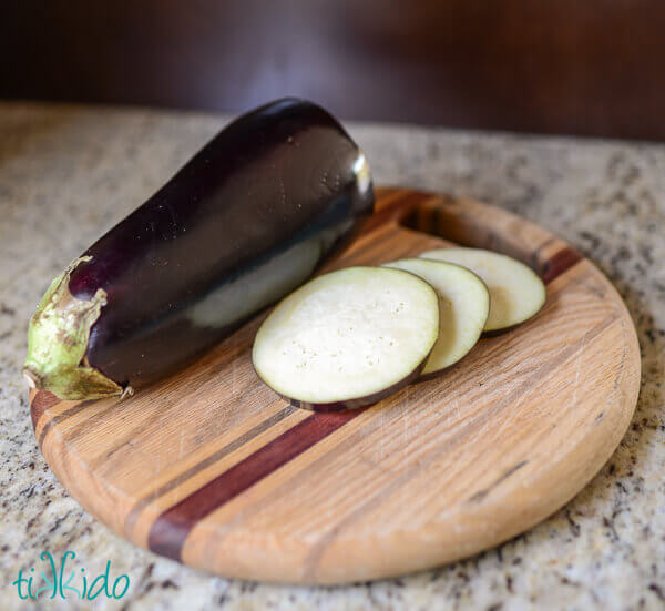Eggplant being sliced on a round wooden cutting board.