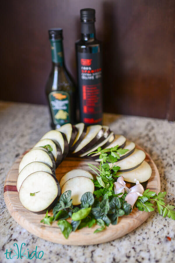 Sliced eggplant and herbs on a round wooden cutting board, olive oil and balsamic vinegar bottles behind.