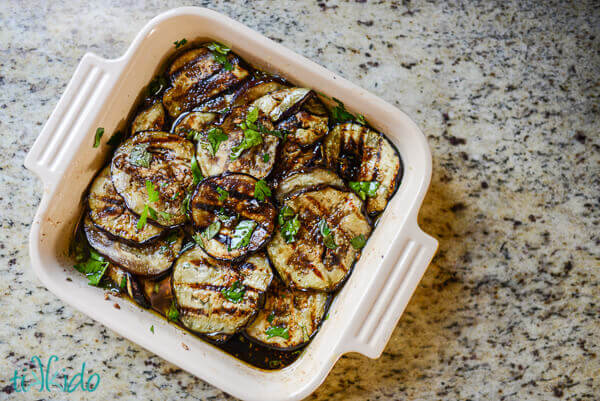 Grilled eggplant slices in a marinade with fresh herbs in a square baking dish on a granite surface.