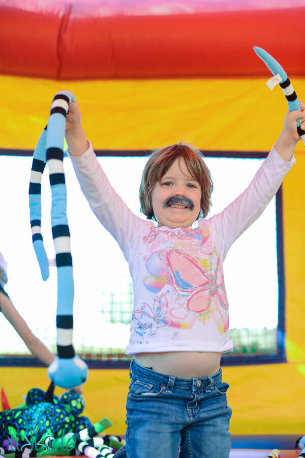 Little girl wearing a fake mustache and holding two stuffed snakes in a bounce house at the Jungle Party