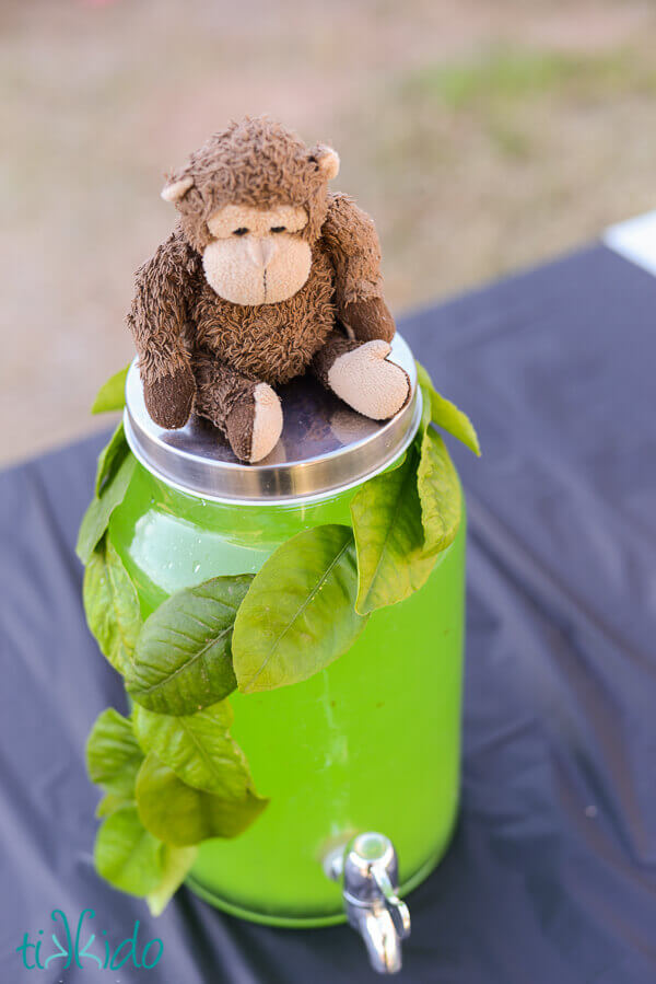Stuffed monkey sitting on a drink dispenser full of green punch and decorated with a garland made from real leaves.