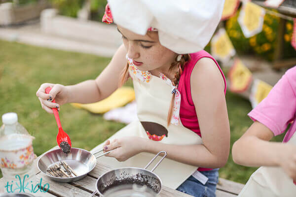 Little girl wearing a chef hat and apron making mud pies.