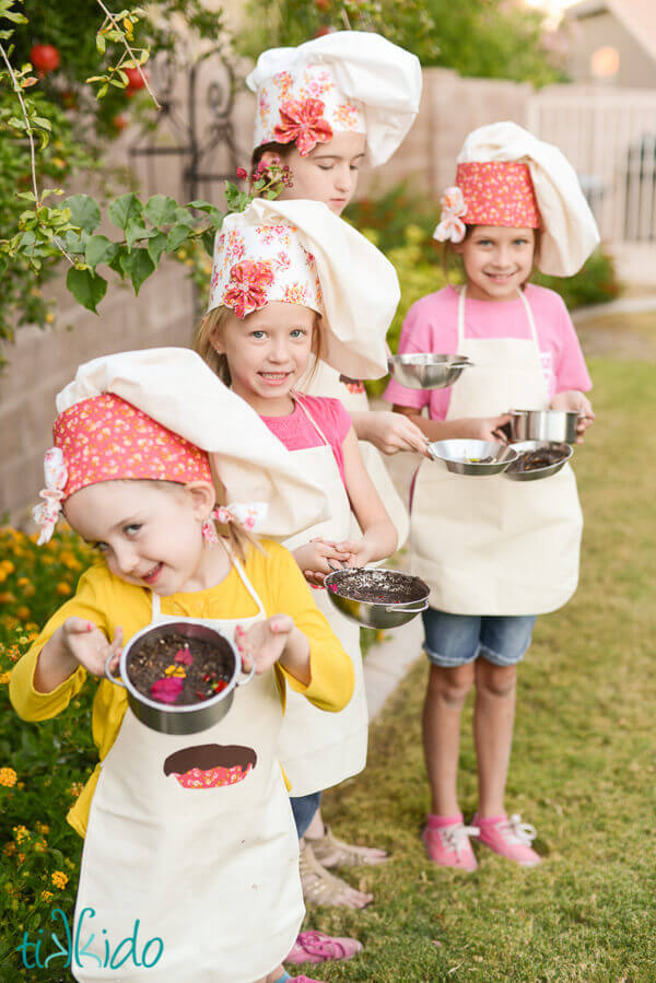 Four little girls wearing chef hats and aprons holding mud pies.