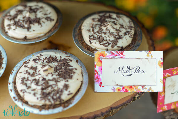 miniature peanut butter cream pie with chocolate graham cracker crust on a rustic tray made of a slice of wood, a tag reading "mud pies" next to pies.