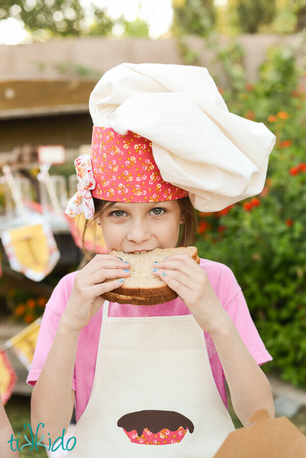 Girl wearing a chef hat and apron eating a sandwich.