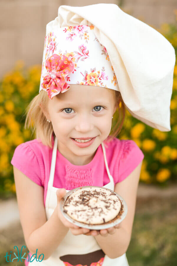 Little girl in chef hat holding Peanut Butter Pie with chocolate graham cracker crust, covered in chocolate shavings.