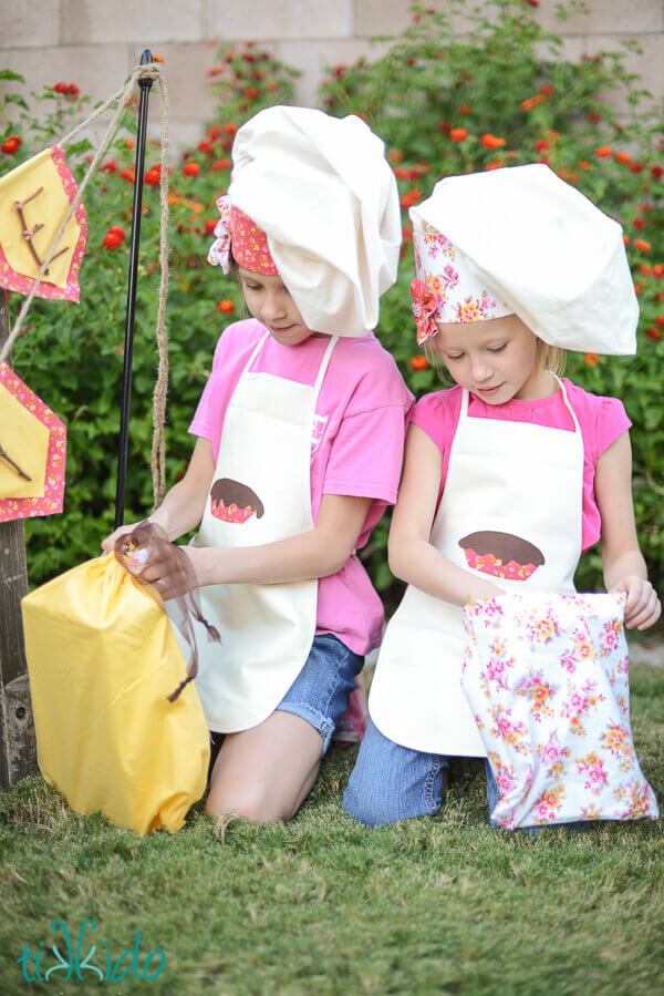 Two little girls wearing chef hats and aprons, opening fabric gift bags at the mud pie birthday party.