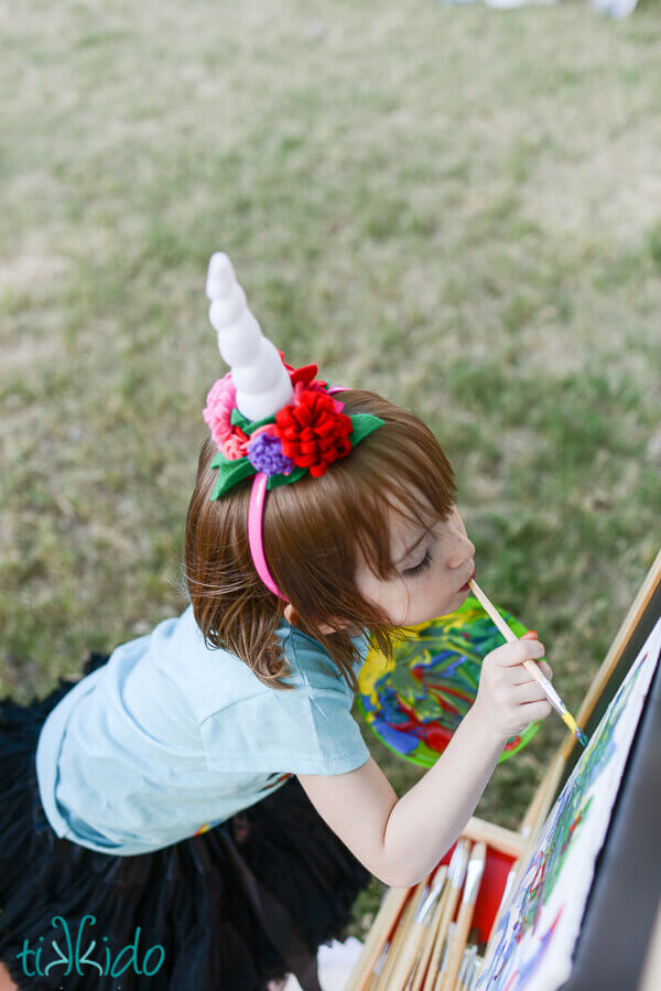 Top view of a little girl wearing a Unicorn horn headband and painting by holding a paintbrush in her mouth.