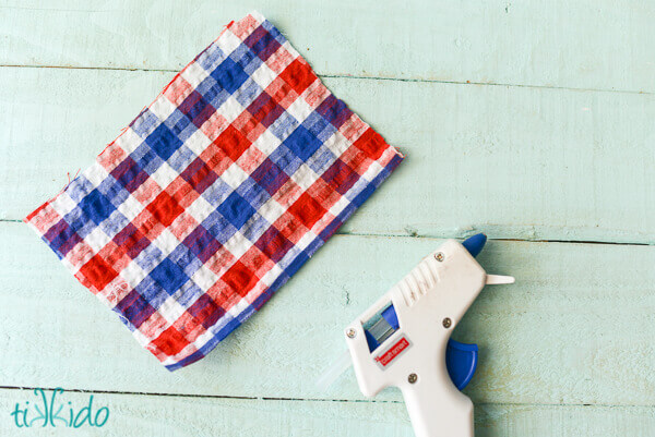 Red, white, and blue seersucker plaid fabric cut into a rectangle next to a glue gun for a no sew bow.