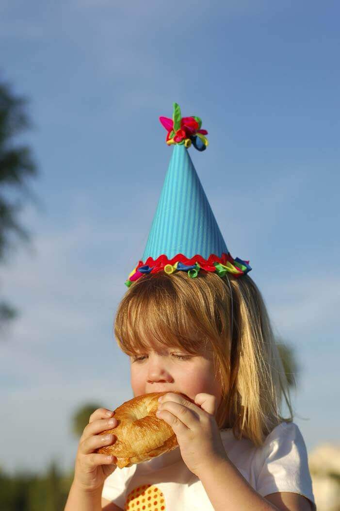 Little girl eating a bagel and wearing a birthday hat decorated with balloons at a balloon themed birthday party.