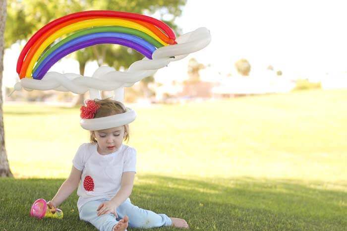 Little girl wearing a large twisted balloon hat that looks like a rainbow and clouds.