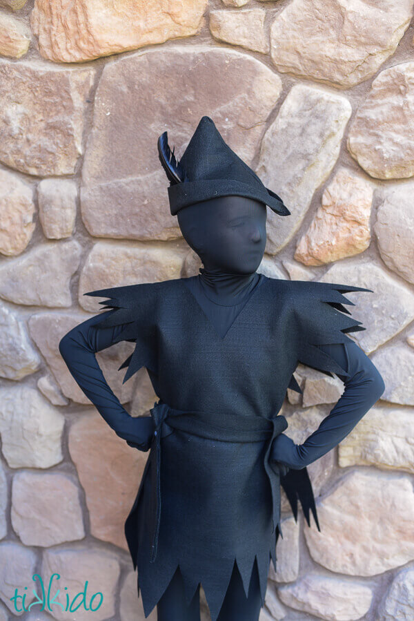 Young girl wearing an all black Peter Pan's shadow costume in front of a stone wall. 